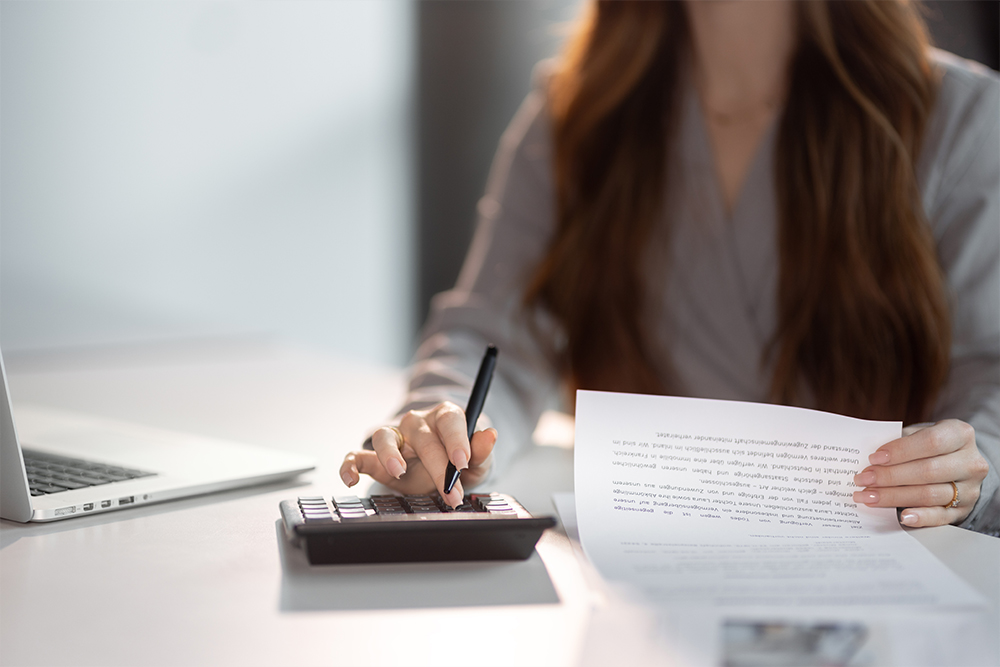 Eine Frau tippt auf einem Taschenrechner und hält ein Dokument in der anderen Hand.
